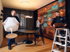 Professionelle Montage bei Freisberg Wohnbedarf: Handwerker installieren eine kunstvolle Wandverkleidung aus Kupferelementen und platzieren Design-Möbel in einem modern gestalteten Raum.